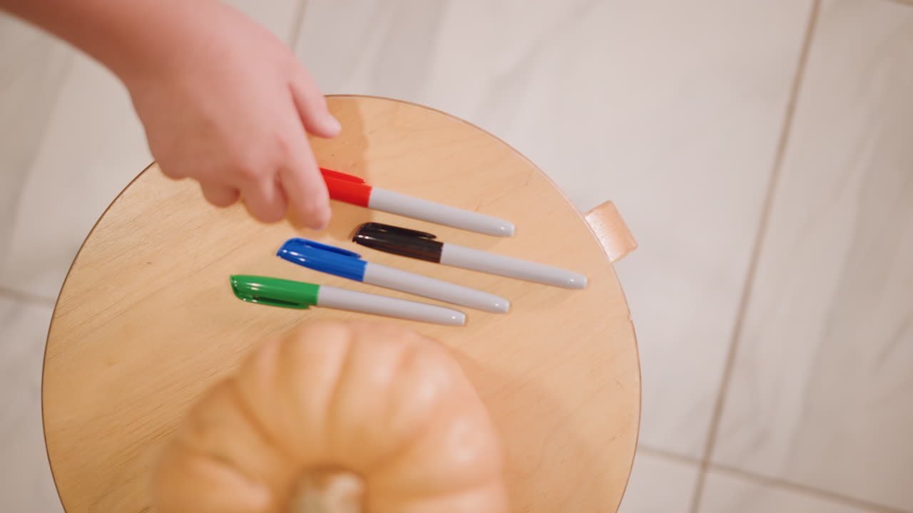 Overhead view pumpkin with markers on round table as hands exchange colors, bright tile floor, collaborative selection for pumpkin design, topdown planning for painting and decoration, playful