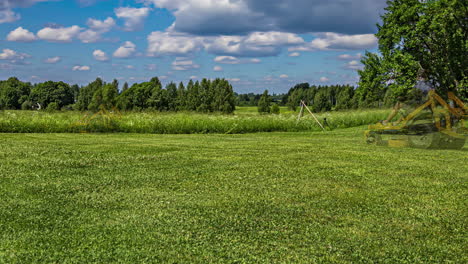 grassy field with trees on horizon under blue sky and cumulus clouds, tractor mowing across grounds