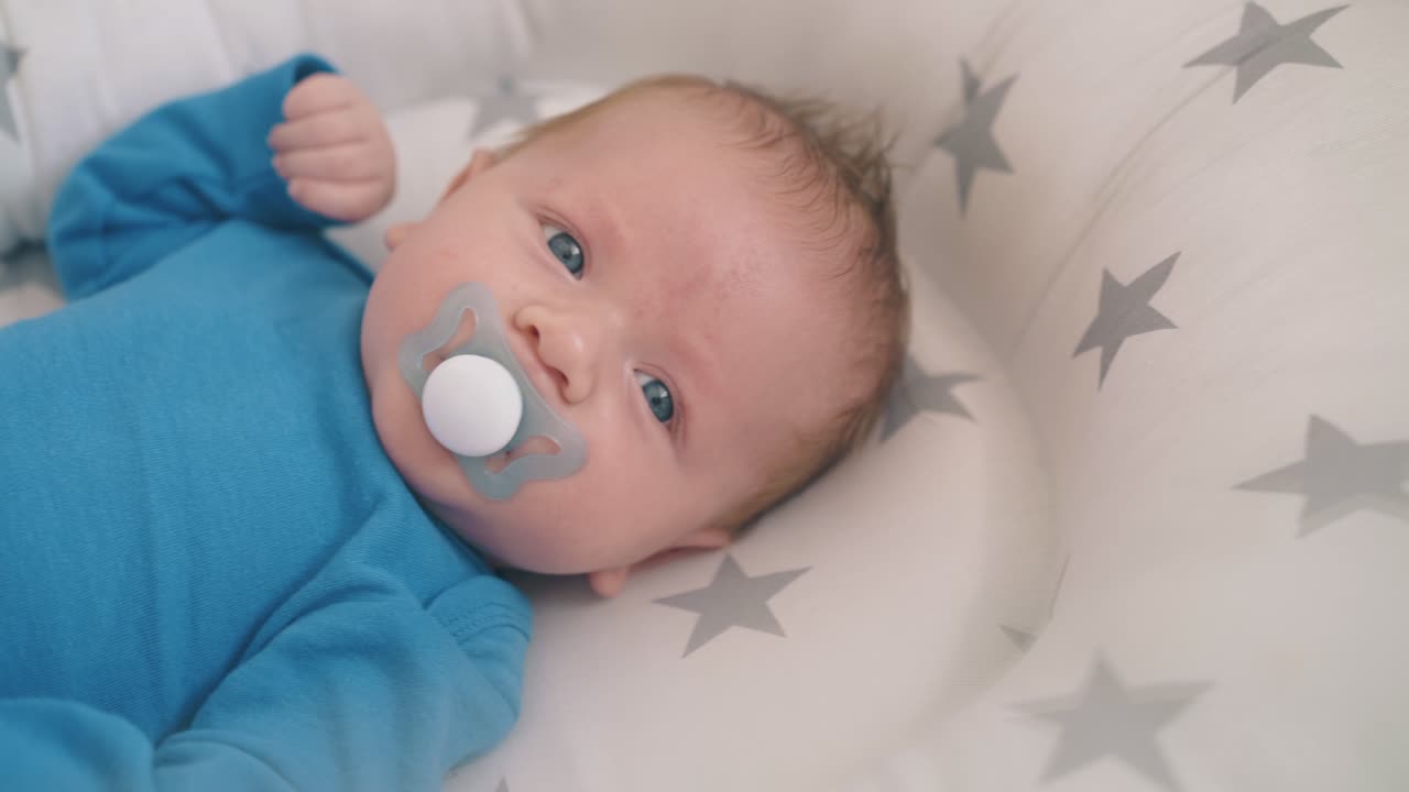 happy newborn baby sucks dummy lying in crib with cocoon