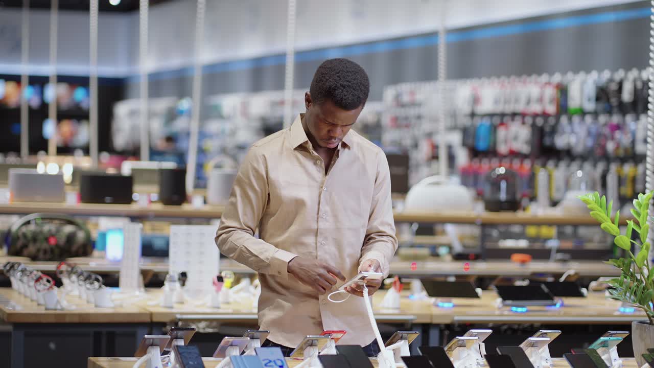 homem afro-americano está escolhendo smartphone em uma loja de equipamentos digitais, retrato de um comprador masculino em um salão de comércio