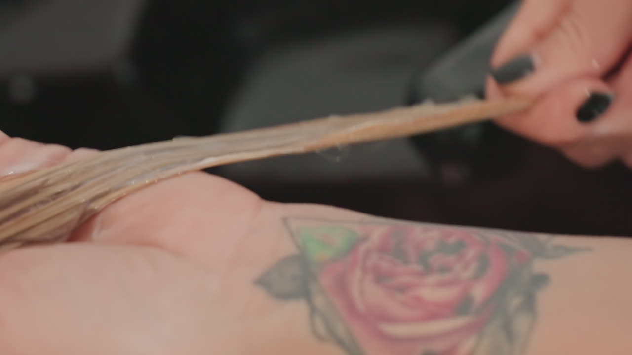 Ciocche di capelli biondi bagnati trattate dalle mani di una stilista non identificata al lavandino del salone; tatuaggio di una rosa sul polso visibile; la stilista divide le ciocche, applica il balsamo, leviga la texture e risciacqua