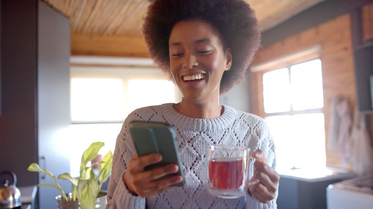 feliz mujer afroamericana usando un teléfono inteligente y bebiendo té en una cocina soleada, en cámara lenta