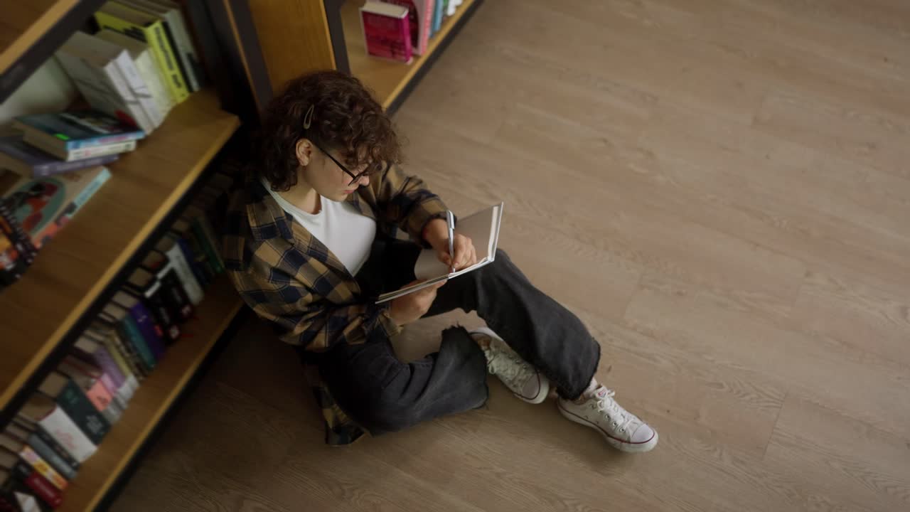 vista superior de una chica con el cabello rizado un estudiante con gafas, leyendo un libro y tomando notas mientras está sentado en el suelo de la biblioteca
