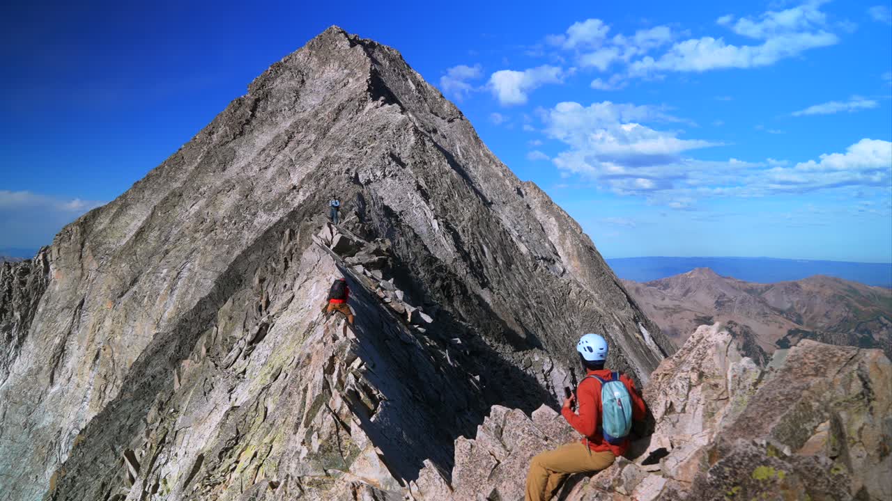 Knifes Edge Capitol Peak north east ridge Wilderness Trail Hikers scrambling rock climbing across Rocky Mountains Colorado 14er mountainside large boudlers aerial view summer blue sky sunny morning