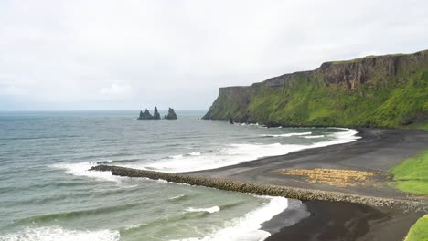 Black-sand-beach-with-large-rocks,-mountains-and-breaker-in-Vik,-Iceland-with-drone-video-moving-down