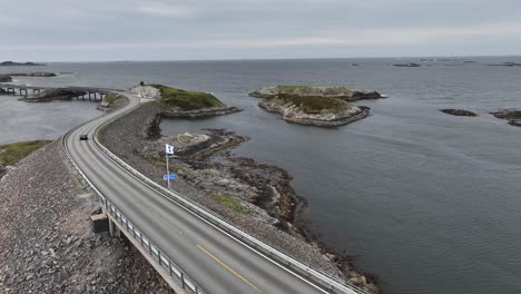 Die-Flagge-Der-Gemeinde-Hustadvika-Weht-Im-Wind-An-Der-Storseisundet-Brücke-Entlang-Der-Berühmten-Atlantikstraße-In-Norwegen---Luftaufnahme-Mit-Darunter-Vorbeifahrendem-Auto-Und-Hintergrund-Des-Atlantischen-Ozeans