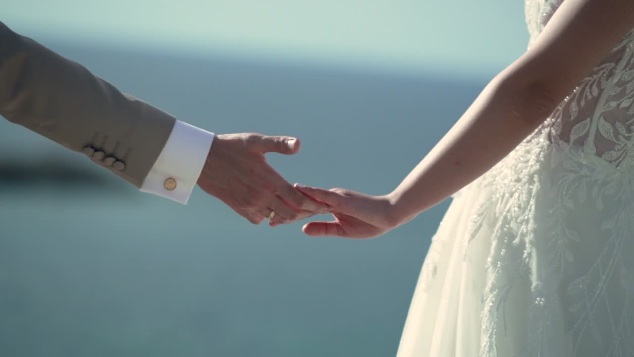 Two individuals hold hands while exchanging wedding rings near the ocean during sunset. The soft light shines on the couple as they share a special moment