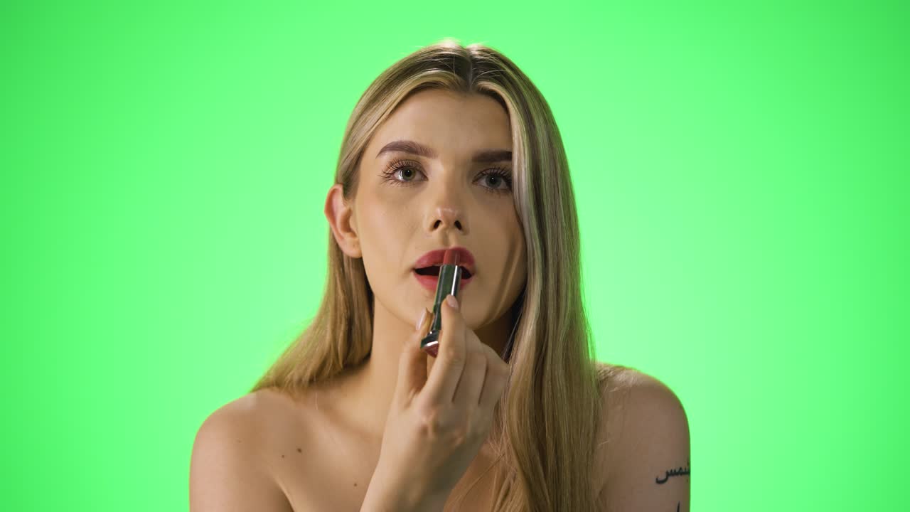 disparo medio de una mujer rubia joven y atractiva mientras aplica lápiz labial rojo en sus labios para prepararse para la noche o la cita mientras se ve sexy ante la cámara frente a un fondo verde en cámara lenta