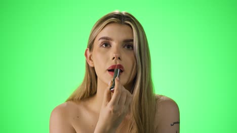 disparo medio de una mujer rubia joven y atractiva mientras aplica lápiz labial rojo en sus labios para prepararse para la noche o la cita mientras se ve sexy ante la cámara frente a un fondo verde en cámara lenta