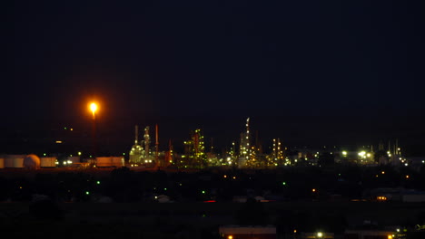 twilight falls on an oil refinery with flaming tower and lights