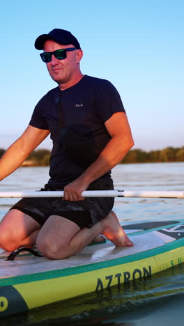 man sits kneeling on the sup board and oaring with one oar. smiling sportsman wearing sunglasses approaches the camera on paddle board. vertical video.