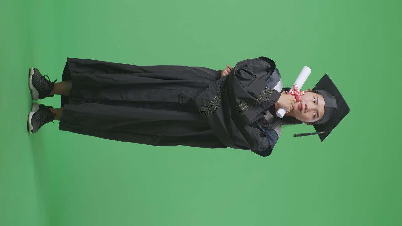 Studentessa asiatica laureata a figura intera con tocco e toga, con il diploma, che pensa e si guarda intorno su sfondo green screen in studio