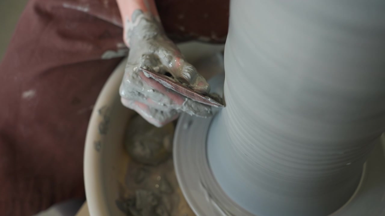 Top view tool trimming tall clay cylinder on spinning wheel, precise scraping motion with rib tool shaping inner wall, wet basin catching excess, energetic workshop rhythm, hands guiding form