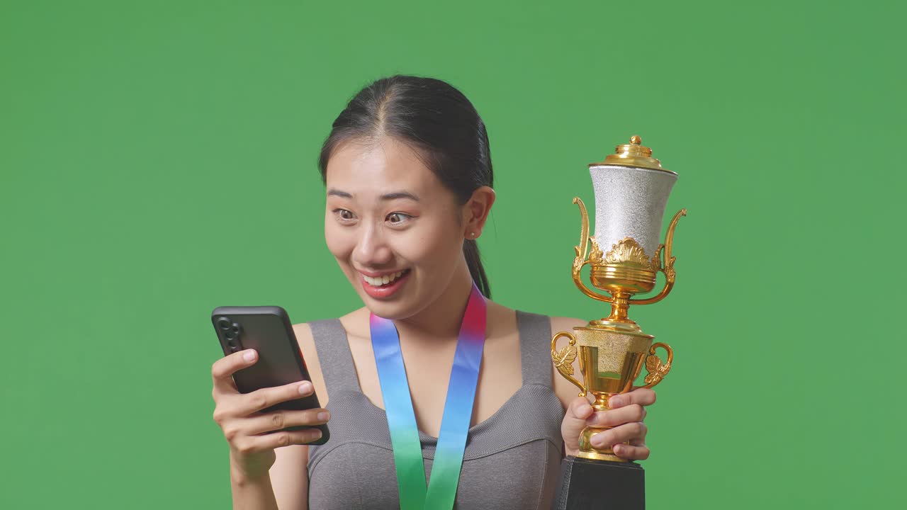 금메달과 트로피를 가진 아시아 여성이 스마트폰을 보고 스튜디오의 녹색 화면 배경에서 와우라고 말하는 클로즈업