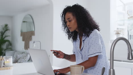 Woman-Wearing-Pyjamas-In-Kitchen-Making-Online-Credit-Card-Purchase-On-Laptop