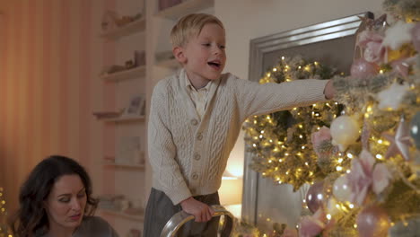 boy decorates christmas tree with balls and ornaments, his mother helps him