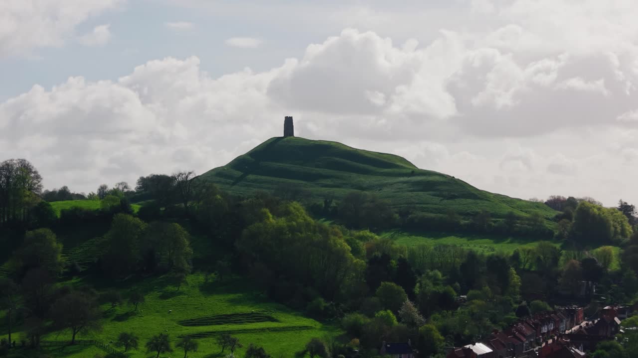 Filmatisk luftfoto af Glastonbury Tor, der rejser sig over frodigt grønt landskab i Somerset, England, med bløde skyer og bølgende bakker, der skaber et dramatisk naturlandskab i Storbritannien.