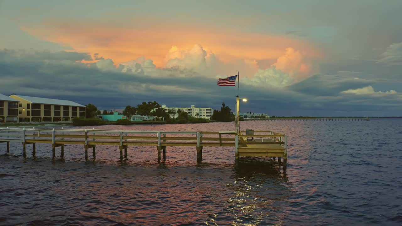 A wooden pier juts into still bay waters beneath a vivid sunset sky, where the American flag waves gently as golden light reflects off the evening horizon