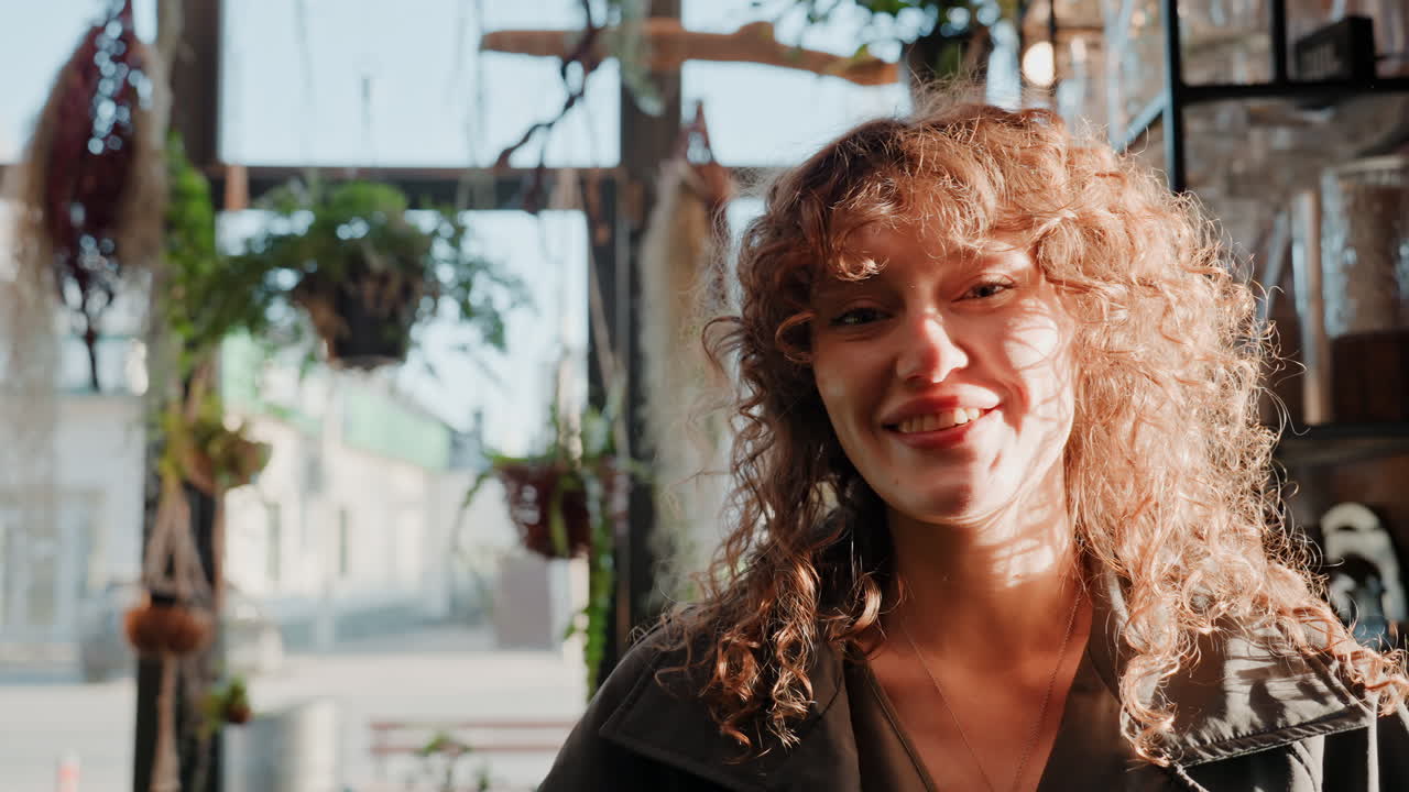 Arrivo alla porta a vetri, fiorista bianca sorridente. Inquadratura di profilo attraverso il vetro d'ingresso con un forte controluce, capelli ricci aureolati dal sole, risata spontanea mentre sistema i vasi, interno intimo con piante pendenti.