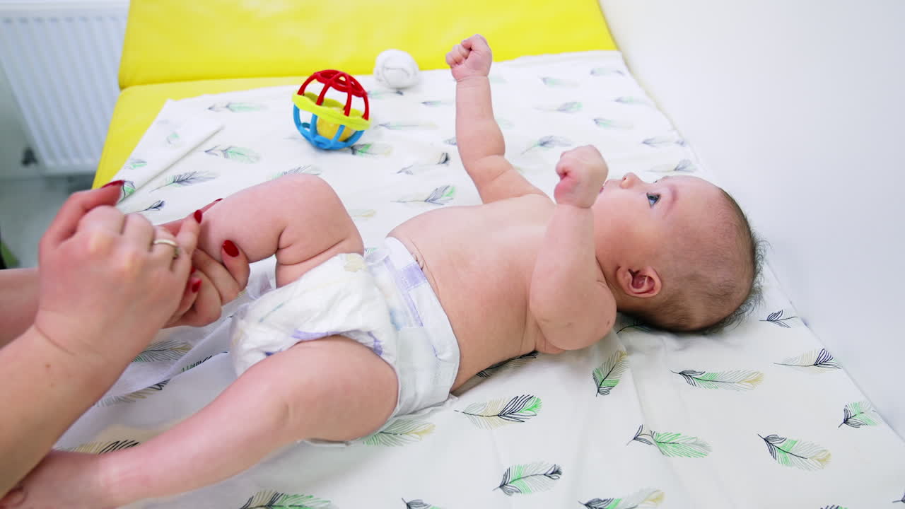 Pediatrician is holding baby's foot in her hands and turning it to check. Toddler at the surgeon's examination. Side view.