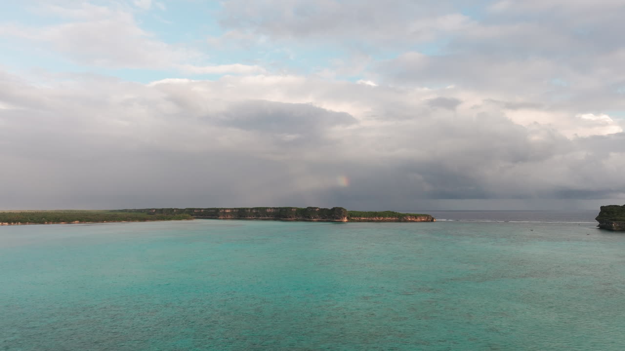 Drone view of Lekini Bay in Ouvéa, New Caledonia, with turquoise lagoon and a faint rainbow over dramatic cloudy sky