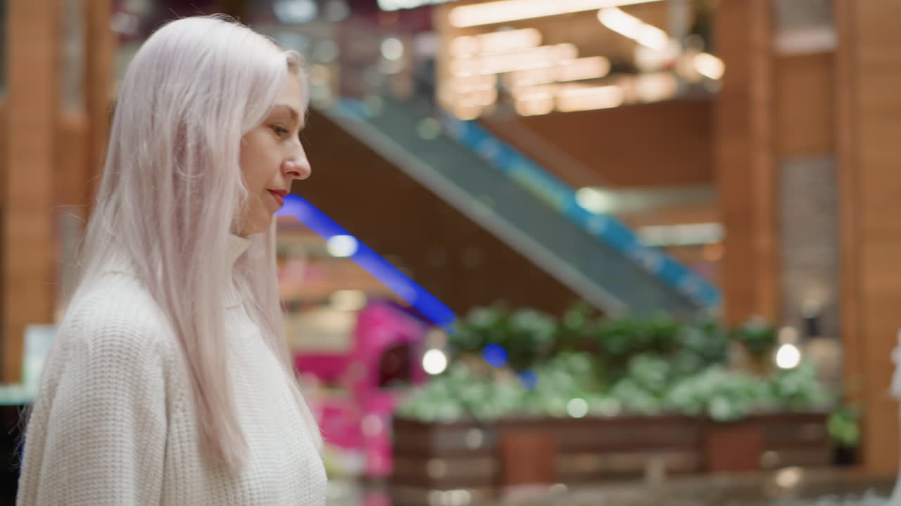 Medium shot of stylish woman walking to illuminated water fountain in mall interior, sitting on tiled bench and gently touching fountain water with fingertips while scanning surroundings