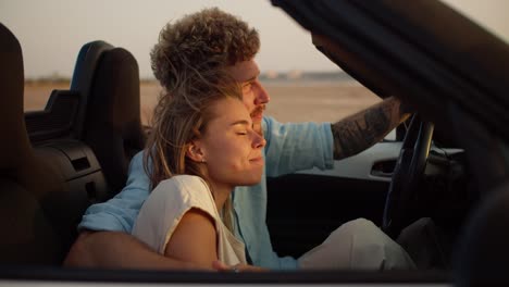 A-bearded-guy-with-curly-hair-hugs-his-blonde-girlfriend-in-a-convertible-car-and-they-look-forward-together-in-windy-weather-on-a-field-background