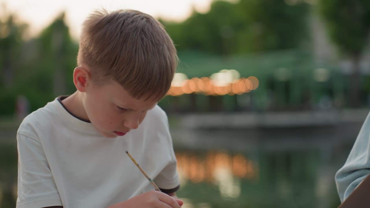 toddler perched on wooden dock beside river under soft sunset glow intently drawing on board with brush, focused expression capturing creative moment during warm evening art session