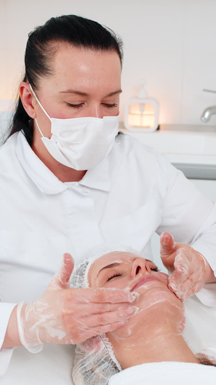 Ahli kecantikan dan kosmetolog memijat serum pengelupas pada wajah wanita dewasa muda paruh baya di klinik