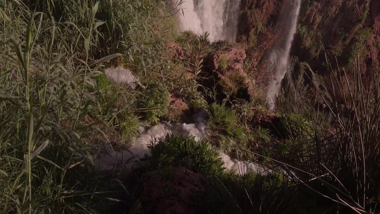Pequeño arroyo fluye rápidamente por hierba alta y rocas rojas junto a la cascada de las Cataratas de Ouzoud.