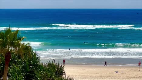 Menschen,-Die-Am-Strand-Mit-Palmen-Entlang-Spazieren,-Mit-Surfern-Im-Hintergrund-An-Der-Goldküste-In-Queensland,-Australien