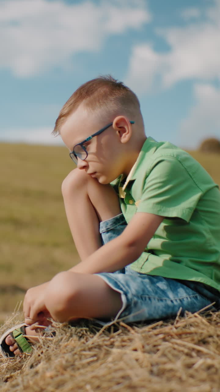 el niño se sienta en una bola de heno con la cabeza inclinada, perdido en sus pensamientos, rodeado de un vasto campo abierto bajo un cielo azul brillante, capturando un momento tranquilo y pacífico en el campo caluroso de un día soleado.