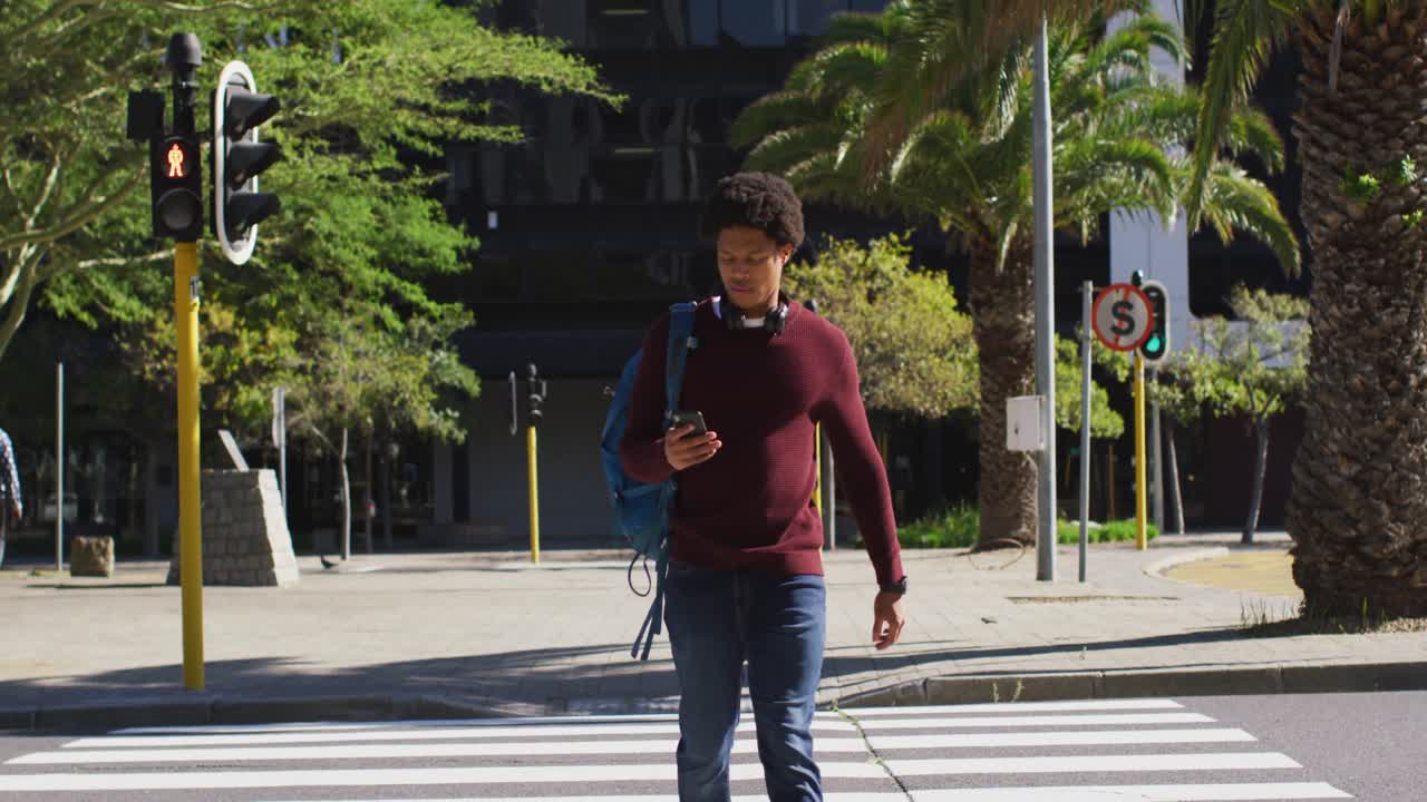 African american man in city, using smartphone, wearing headphones and backpack crossing street