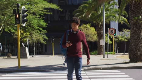 african american man in city, using smartphone, wearing headphones and backpack crossing street