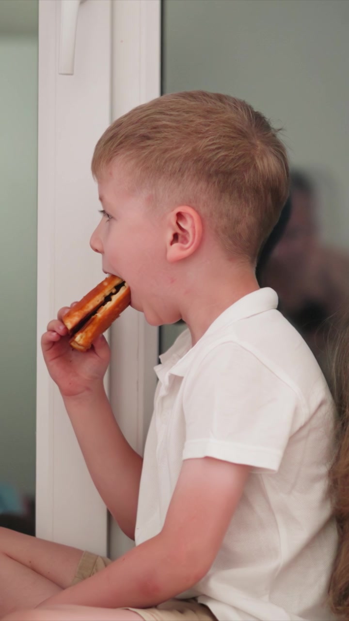 Close up of young boy eating waffle while sitting near window. Boy holds and takes bites from food item with another boy partially visible beside him. Soft lighting creates natural indoor