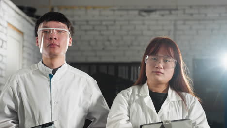 male technician nods warmly while holding tablet, as colleague adjusts safety glasses and tilts head slightly with a soft smile, white light glows behind her in automotive workshop setting