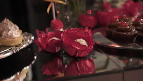 two vibrant red paper flowers resting on a reflective surface, enhancing a dessert table adorned with an array of pastries and sweets, creating an elegant and festive atmosphere, slow motion reveal