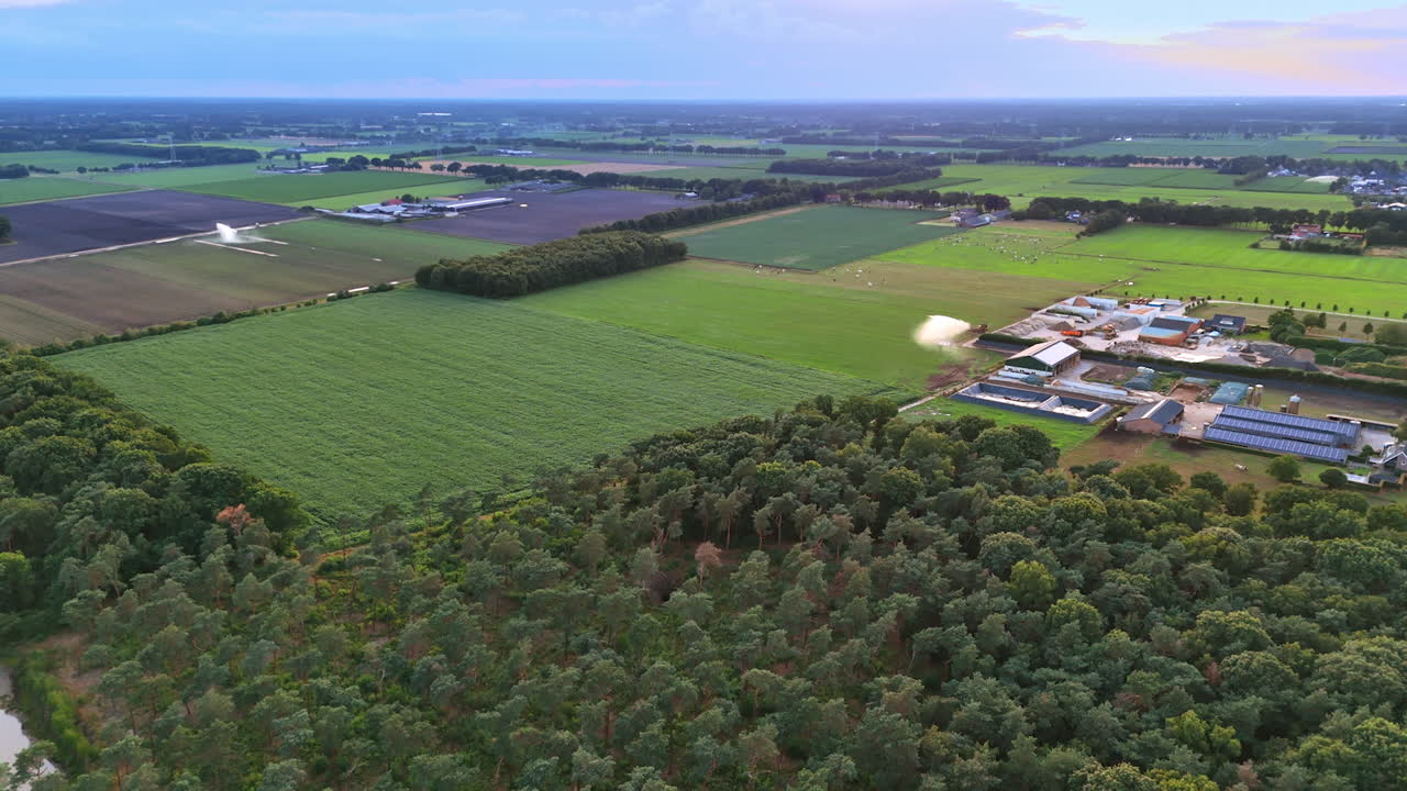 Farmland and forest from above. Vast green fields stretch across the land, bordered by dense trees and agricultural buildings during twilight