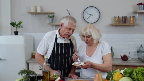 senior couple in kitchen. grandmother feeding grandfather with raw sprouts buckwheat with nuts