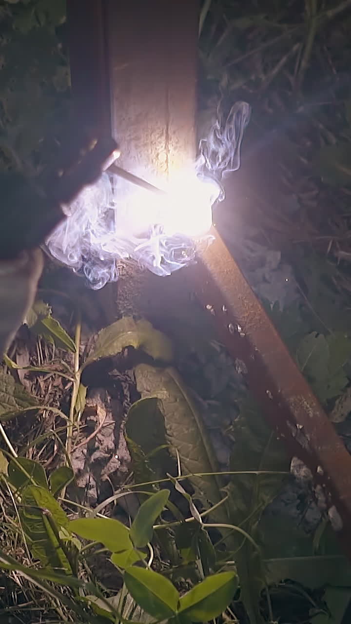 slow motion closeup worker in dirty protective gloves welds plank to metal fence pole in dark garden