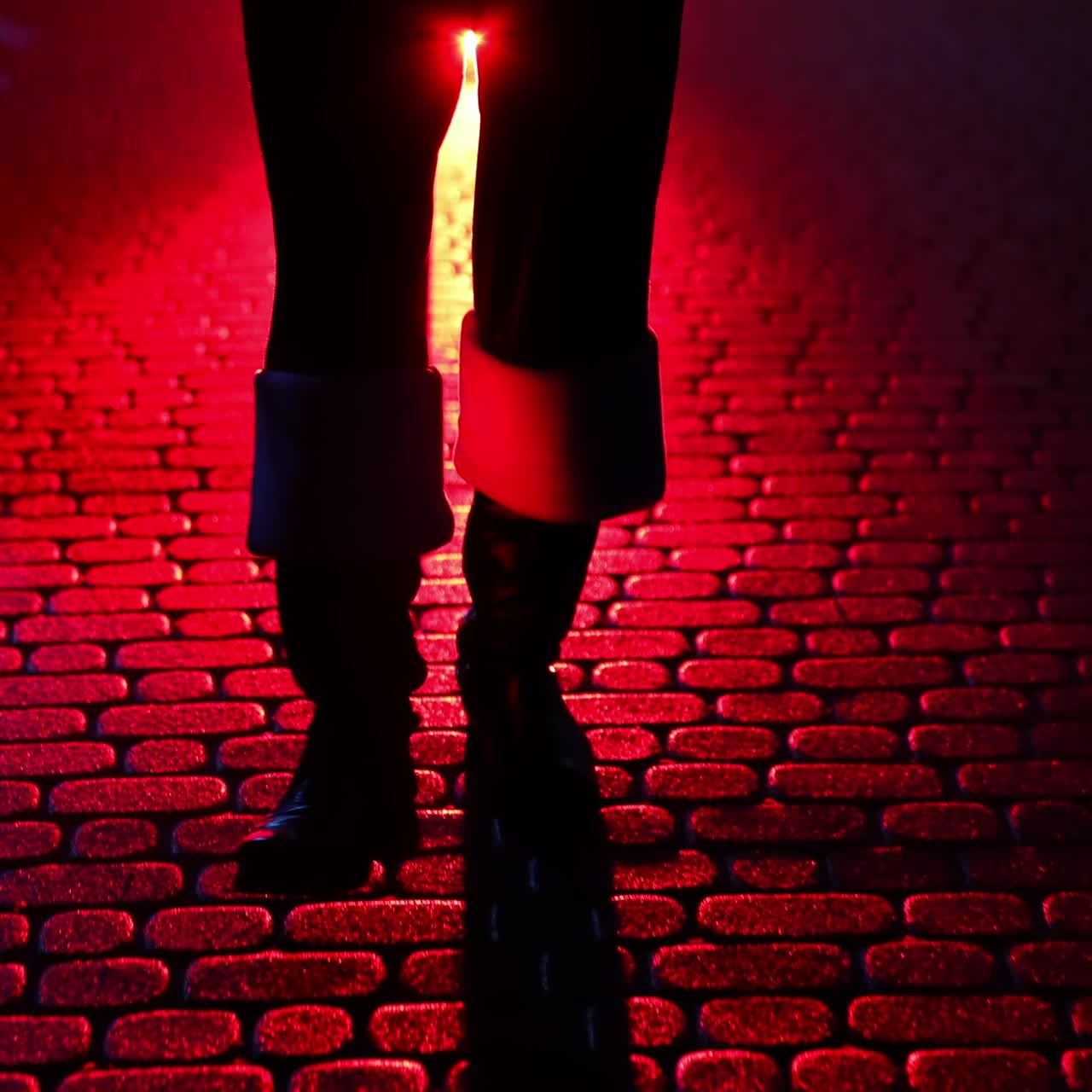 Legs of Santa Claus. Father Frost carrying presents at Christmas in red bag outdoors. Black boots of Santa walking on a pavement at dark on snow background. Red light