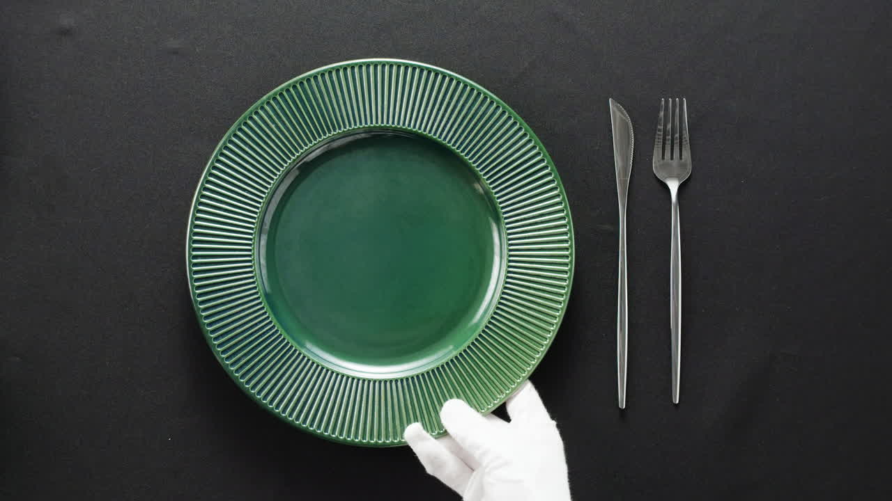 Minimalist table setting with white plate, fork, and knife arranged neatly, and a gloved hand adjusting cutlery.