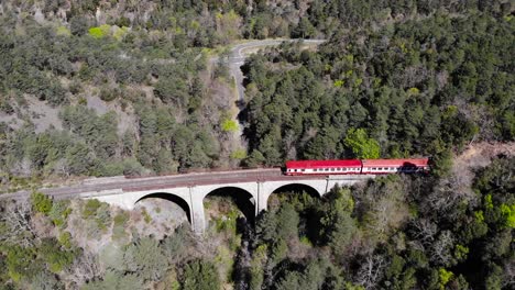 Aerial:-red-and-white-old-train-in-a-forest-crossing-an-ancient-viaduct-in-southern-France