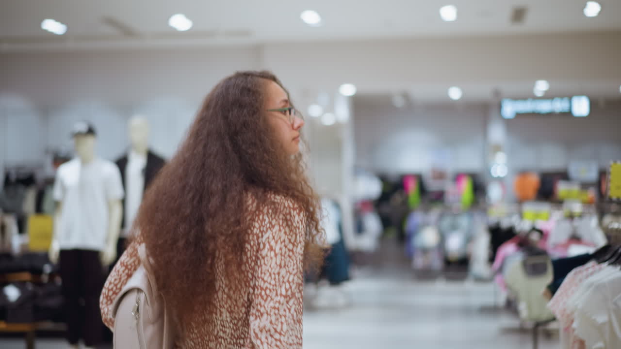 한 여성이 그녀의 가방과 함께 밝게 밝은 옷 가게를 탐험하고, 주변을 둘러보고 랙에 있는 물건을 만지며, 세련된 옷과 잘 조직된 공간에 둘러싸인 캐주얼 쇼핑 경험을 포착합니다.