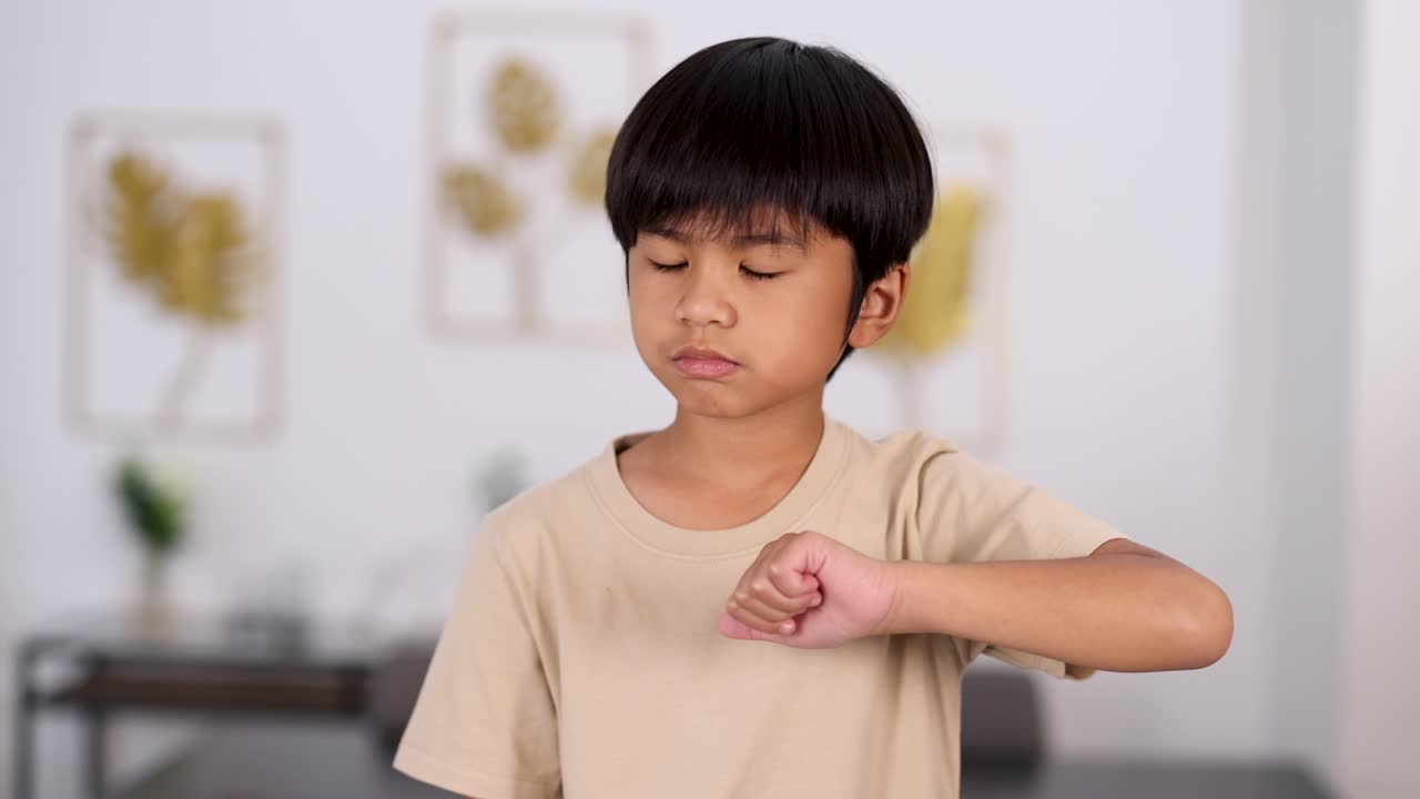 Young Asian boy in beige shirt performs thumbs down gesture with serious expression in a bright, modern room. Soft, natural lighting and static camera