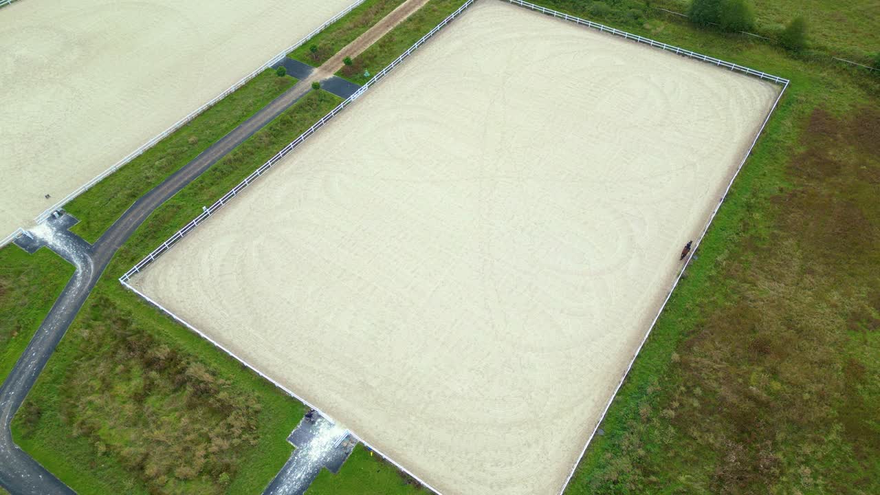 Aerial view of horseback riding hall with solar panes on roof and outside arena with two people on horseback