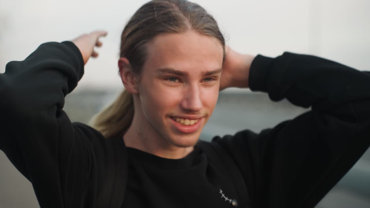 Smiling young man tying hair, relaxed grin on overpass at dusk, casual sweater and backpack, hands gather ponytail after braids, candid joyful moment with warm soft light and urban background