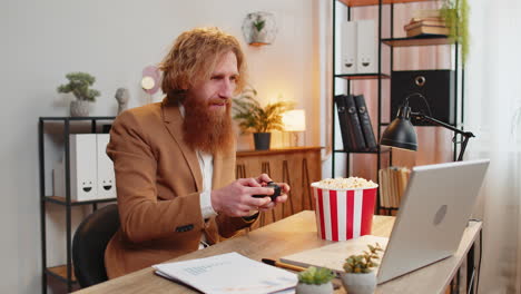 businessman eating popcorn while playing video game on laptop, spending leisure time at home office