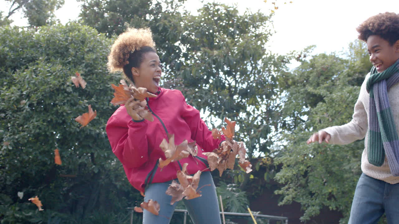 Happy african american mother and son in warm clothes throwing leaves in garden, slow motion