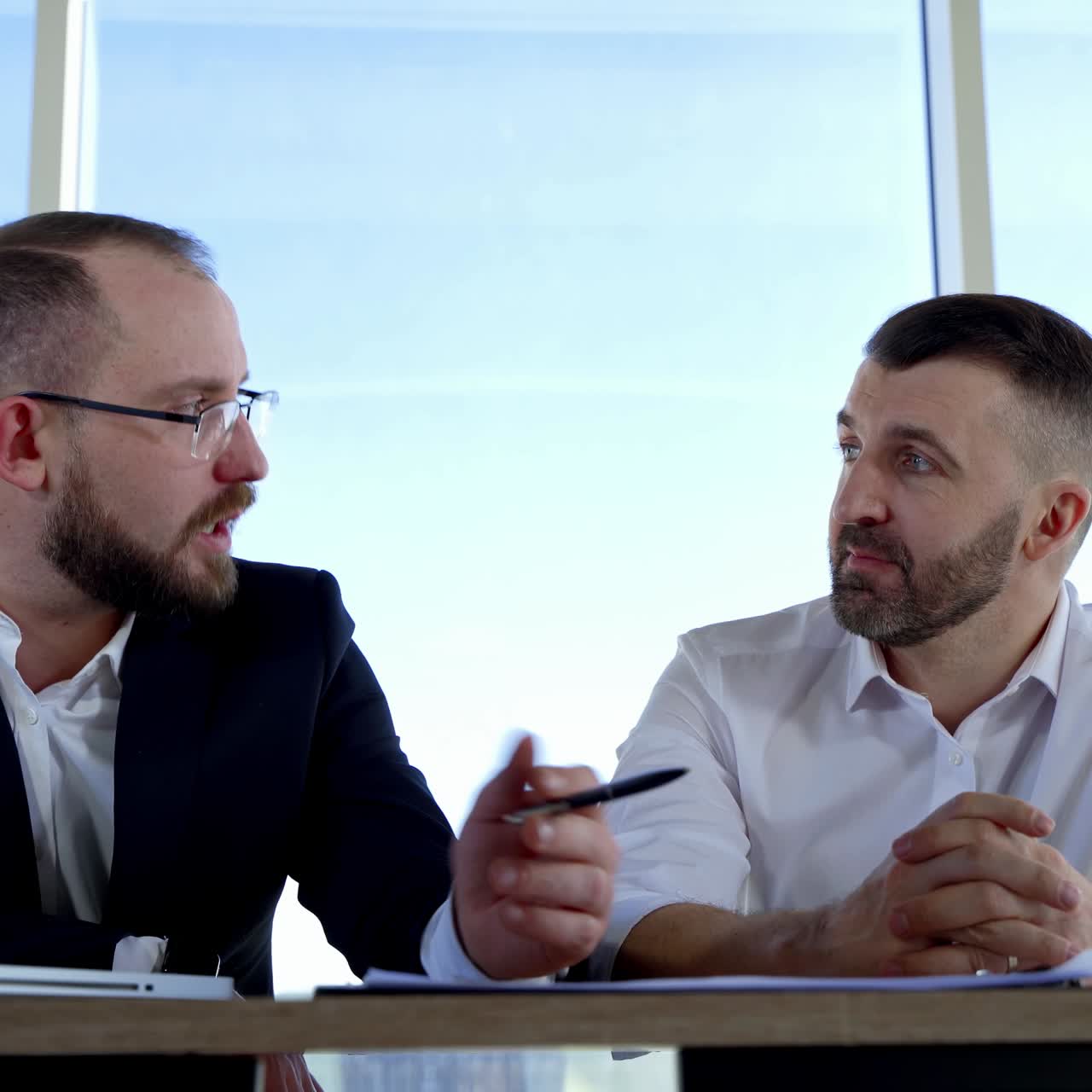 Bearded business partners talking in office. Male office workers having a conversation about business while sitting at the table by the window